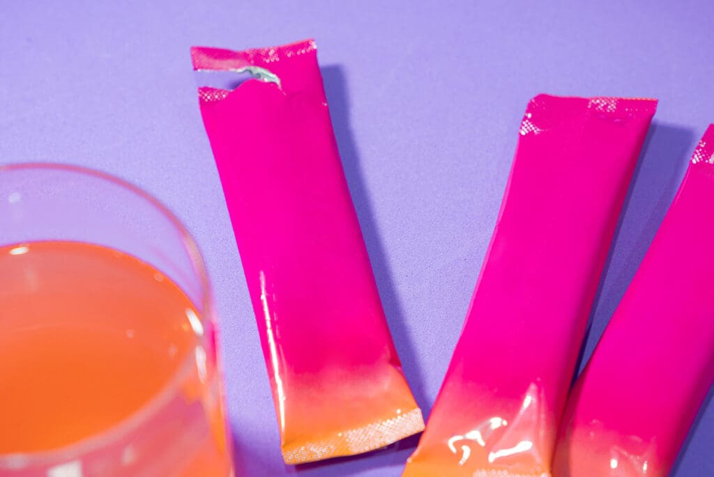 Colorful drink mix packets with glass on purple background.