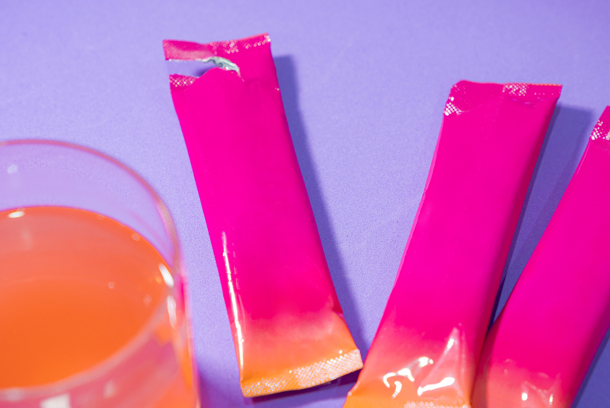 Colorful drink mix packets with glass on purple background.
